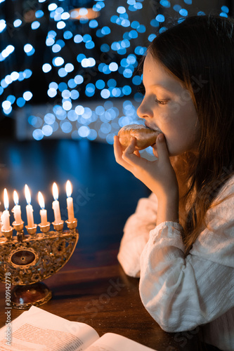 Child girl eating sufganiyot donut with Menorah Candles on wooden table and sufganiyot on background light glitter bokeh overlay. Hanukkah jewish holiday Israel hebrew traditional family celebration photo