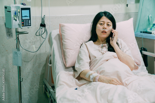 unhappy young Asian chinese female patient talking on mobile phone in bed. attractive sick woman lying on bed in hospital room. illness people connected world and mobile office concept.