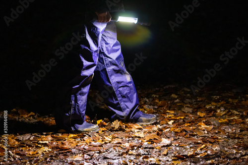 Kind läuft mit Taschenlampe durch dunkle Nacht im Wald mit blauer Regenhose und Gummistiefeln auf feuchtem Waldboden mit buntem Laub allein und voller Angst als Nachtwanderung