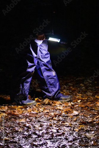 Kind läuft mit Taschenlampe durch dunkle Nacht im Wald mit blauer Regenhose und Gummistiefeln auf feuchtem Waldboden mit buntem Laub allein und voller Angst als Nachtwanderung photo