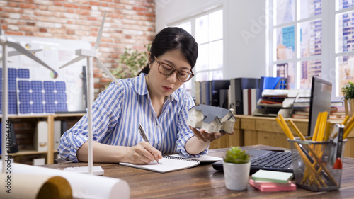 pretty female architect working on notebook. asian korean woman engineer in workplace planning architectural project with green energy. elegant girl worker writing new ideas note with 3D house model