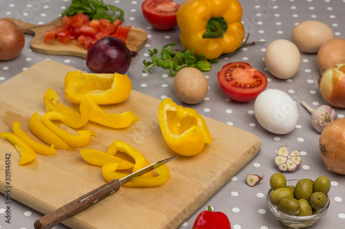 Slicing yellow pepper on a cutting board. Eggs, olives, garlic on the table..
