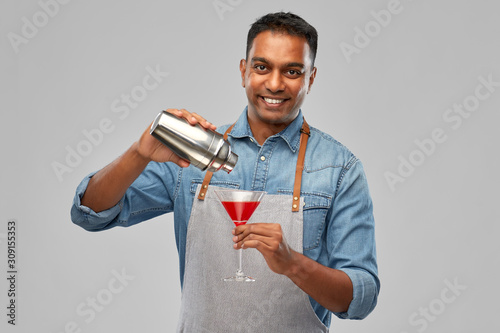alcohol drinks, people and profession concept - indian barman in apron pouring cocktail from shaker to glass over grey background