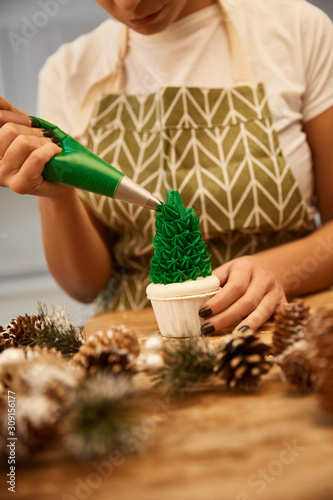 Cropped view of confectioner pouring sweet green cream on Christmas tree cupcake beside spruce cones on table