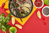 top view of pho in bowl with chopsticks near sauces, lime, chili and coriander on red and yellow background