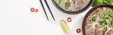 top view of pho in bowl near chopsticks, lime, chili and soy sauces and coriander on white background, panoramic shot