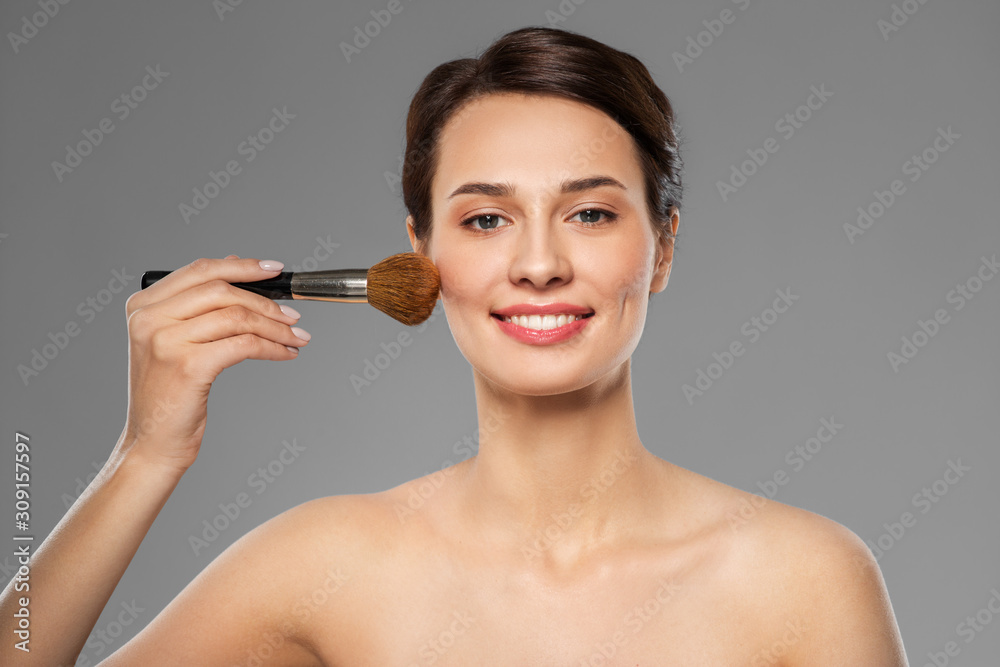 Fototapeta premium cosmetics and beauty concept - beautiful woman with makeup brush applying blusher over grey background