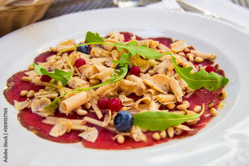Thinly sliced deer carpaccio with pine nuts, truffles and blueberries, served on white plate, decorated. Delicious food in a typical popular restaurant in Riga, Latvia Thinly sliced deer carpaccio with pine nuts, truffles and blueberries, served on white plate, decorated. Delicious food in a typical popular restaurant in Riga, Latvia
