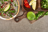 top view of pho in bowl near lime, chili and coriander on wooden cutting board on grey background