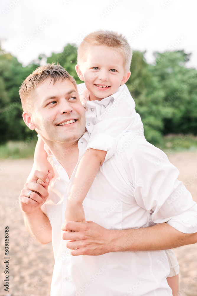 Fototapeta premium Happy dad carries on the back of his smiling son. Father and son walk on the beach, dressed in bright clothes. Vertical format.