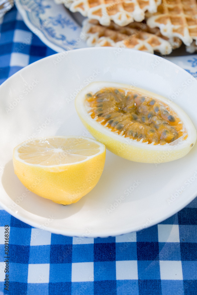Picnic table decoration on nature with a white plate with lemon and passion fruit and waffles, everything stands on a blue checkered tablecloth. Picnic table decoration on nature with a white plate with lemon and passion fruit and waffles, everything stands on a blue checkered tablecloth.