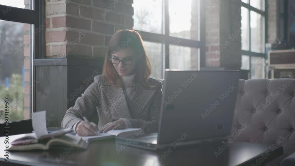 distance learning, successful business woman with glasses is studying online making notes in a notebook while working on a laptop computer in a cafe