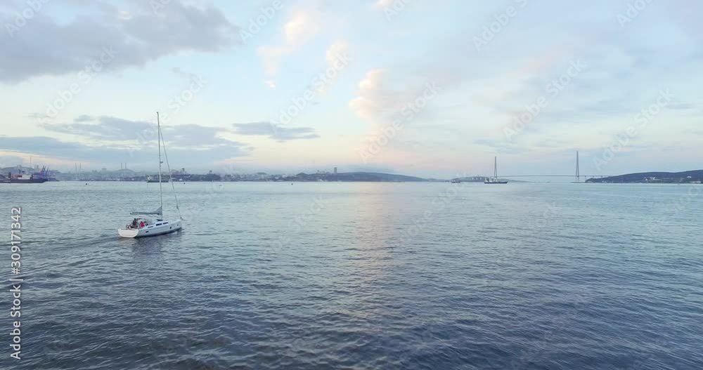 Aerial view of bautiful white sailing yacht with high mast in the Eastern Bosphorus strait at summer sunset. Vladivostok cityscape and Russian bridge are on the background Aerial view of bautiful white sailing yacht with high mast in the Eastern Bosphorus strait at summer sunset. Vladivostok cityscape and Russian bridge are on the background