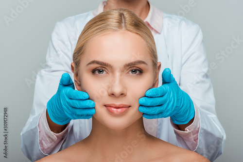 cropped view of beautician in blue latex gloves touching cheeks of woman isolated on grey