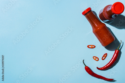 Top view of bottles with spicy tomato sauce beside chili peppers on blue background