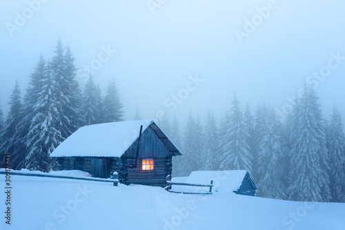 Fantastic winter landscape with wooden house in snowy and foggy mountains forest. Christmas holiday and winter vacations concept