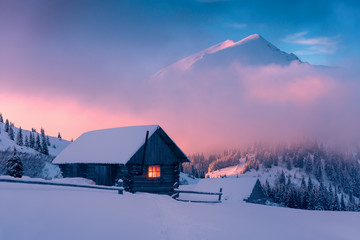 Fantastic winter landscape with wooden house in snowy mountains. Hight mountain peaks in foggy sunset sky. Christmas and winter vacations holiday concept