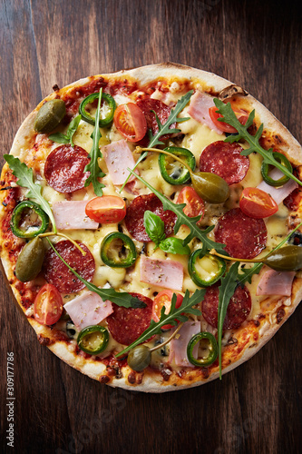 Pizza with salami, ham, cherry tomatoes, jalapeno pepper and rocket. Home made food. Concept for a tasty and hearty meal. Brown wooden background. Top view. 