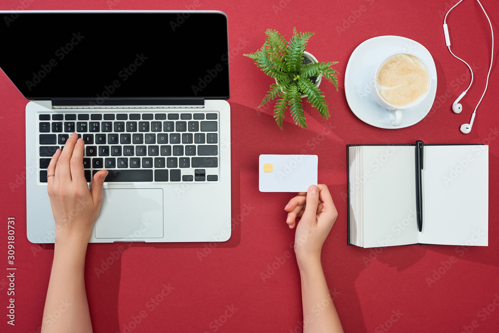 Obraz premium cropped view of woman holding credit card and using laptop near earphones, coffee, notebook with pen and plant on red background