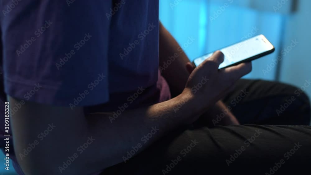 Young man wearing violet t-shirt and watch is sitting in room with dimmed light and holding tablet in hands, actively using it , while camera is automatically moving around him. Rotation video.