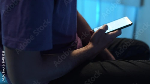 Young man wearing violet t-shirt and watch is sitting in room with dimmed light and holding tablet in hands, actively using it , while camera is automatically moving around him. Rotation video.