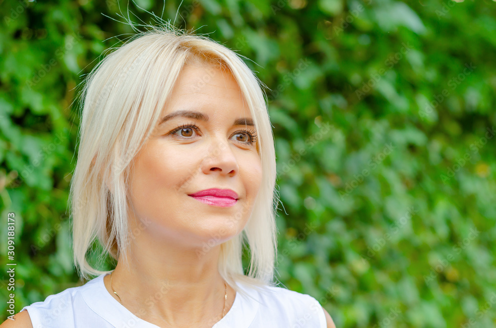 Beautiful blonde middle-aged woman in green leaves background. In white blouse, standing on street. Pretty smile on face straight short hair, brown eyes Beautiful blonde middle-aged woman in green leaves background. In white blouse, standing on street. Pretty smile on face straight short hair, brown eyes