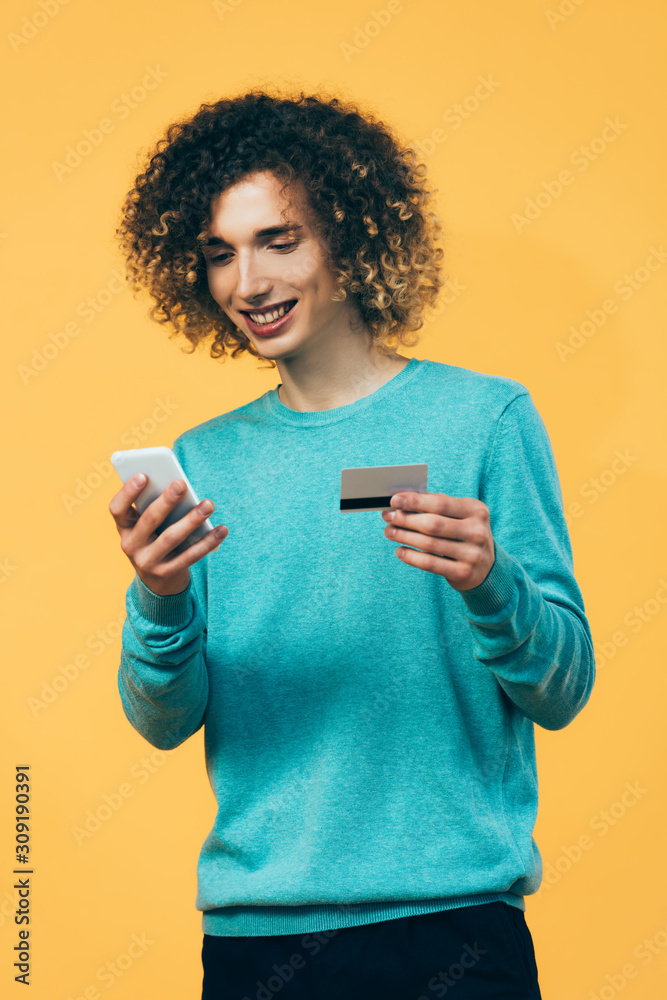 smiling curly teenager holding smartphone and credit card isolated on yellow smiling curly teenager holding smartphone and credit card isolated on yellow