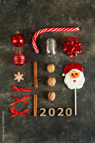 Christmas decor are laid out on a dark concrete surface in the shape of a rectangle: Santa Claus lollipop, Christmas cane, nuts, red bows, Christmas balls, cinnamon sticks, present and gingerbread