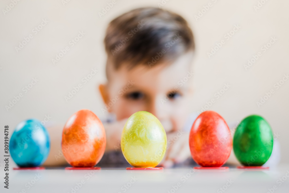 Fototapeta premium Multi-colored Easter eggs stand in a row on stands on the background of a boy looking at them.