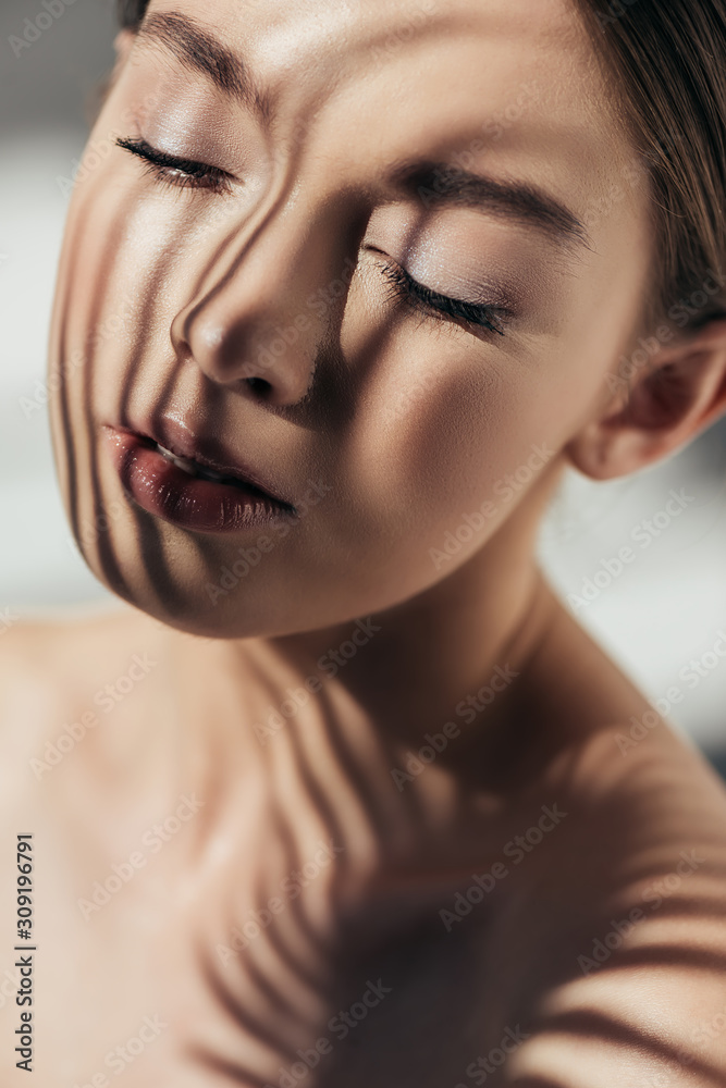 Naklejka premium portrait of naked girl with closed eyes and shadows on face on grey