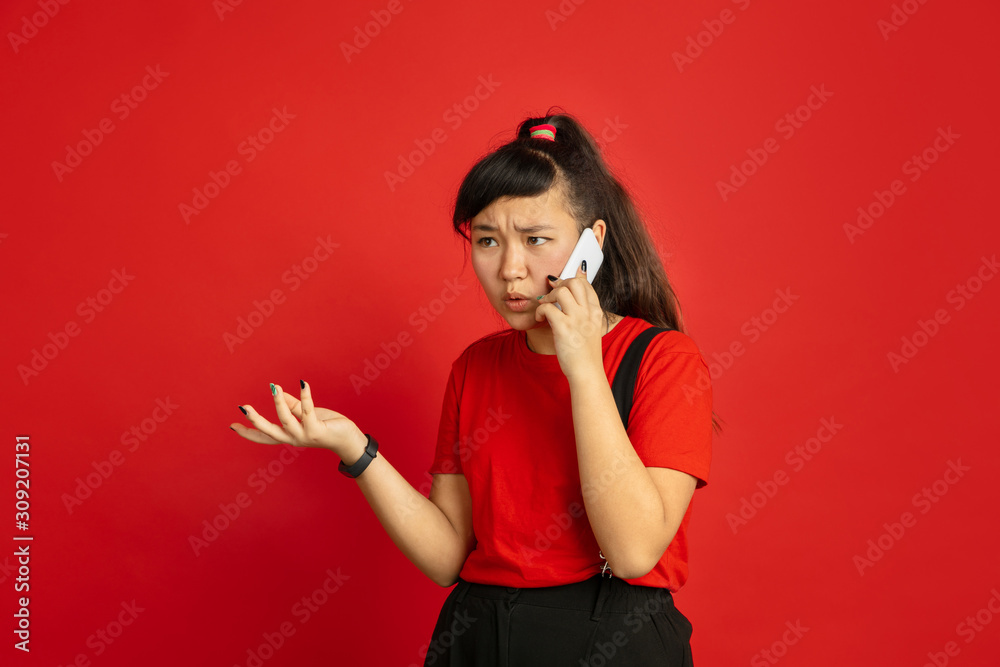 Asian teenager's portrait isolated on red studio background. Beautiful female brunette model with long hair in casual style. Concept of human emotions, facial expression, sales, ad. Talking on phone. Asian teenager's portrait isolated on red studio background. Beautiful female brunette model with long hair in casual style. Concept of human emotions, facial expression, sales, ad. Talking on phone.