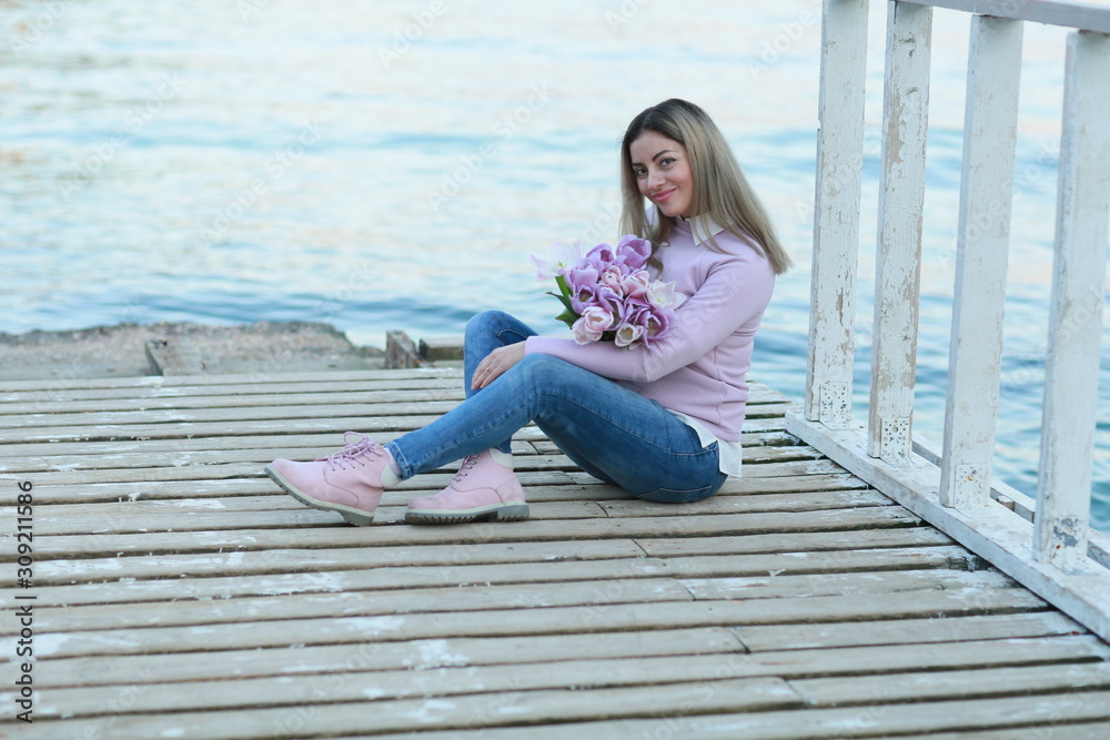 A woman with flowers in her hands sits on a wooden pier near the water on the background of the mountain.