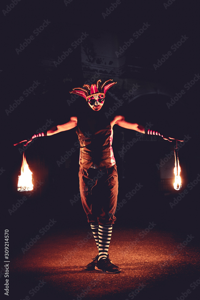 Man in red jester costume with holding double fire chains making some action moment, standing in the dark - obrazy, fototapety, plakaty 