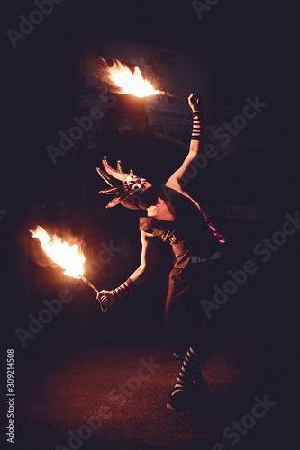 Man in red jester costume with double fire torch performing some action moment in the dark, toning style