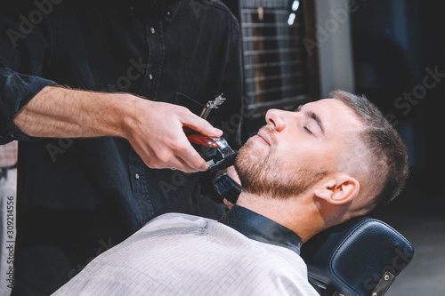 Young male barber shaves the client's beard with a electric trimmer. Young bearded man getting haircut by hairdresser at barber shop. Advertising and barber shop concept