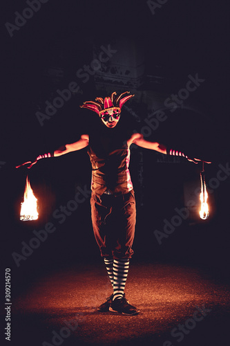 Man in red jester costume with holding double fire chains making some action moment, standing in the dark