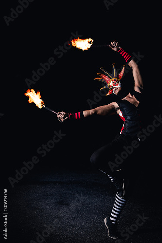 Man in red jester costume with double fire torch performing some action moment stand with one leg in the dark