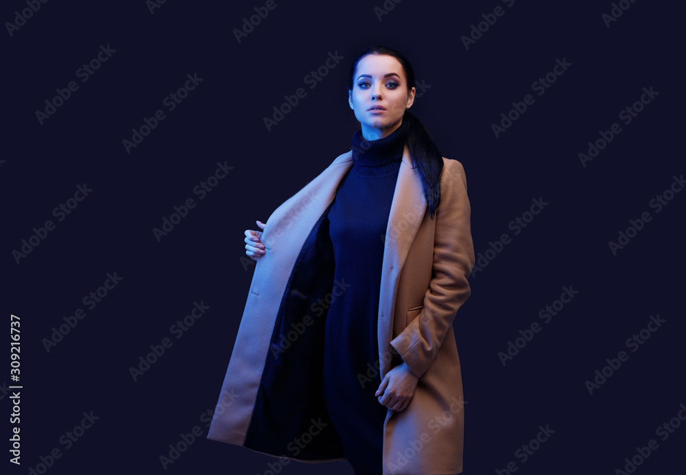 Beautiful woman on dark background. Portrait of charming lady in beige classic coat and black dress. Blue neon light. Free space for text. Beautiful woman on dark background. Portrait of charming lady in beige classic coat and black dress. Blue neon light. Free space for text.