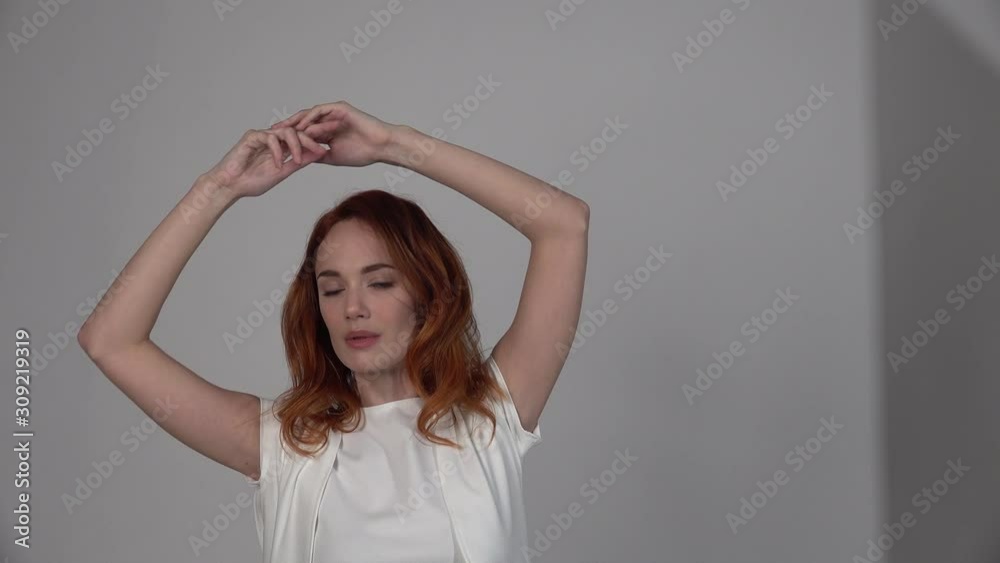 Beautiful redhead girl in a light beige dress demonstrates a model pose on an isolated neutral grey background. Fashion, modern clothes and style, Graceful dance movement of the arms above the head. Beautiful redhead girl in a light beige dress demonstrates a model pose on an isolated neutral grey background. Fashion, modern clothes and style, Graceful dance movement of the arms above the head.