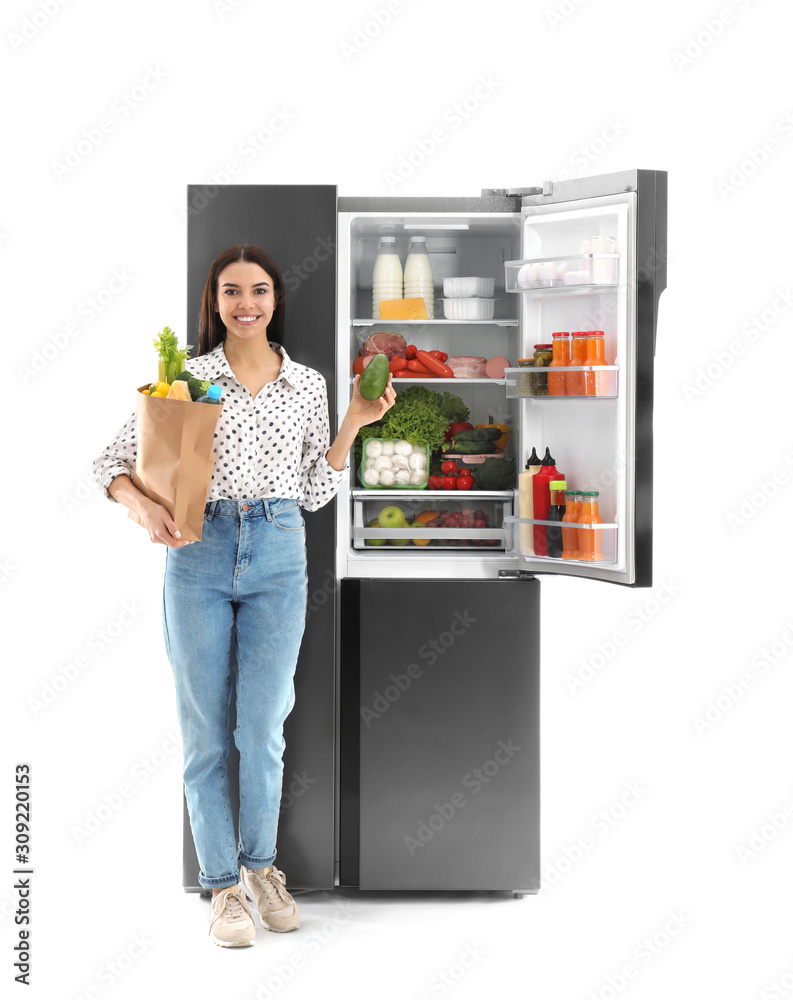 Young woman with bag of groceries near open refrigerator on white background