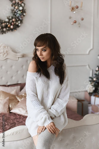 Young beautiful woman with dark hair in fashionable cozy clothes posing in decorated for Christmas bedroom. Sexy model girl with perfect body in knitted dress and stockings at New years interior