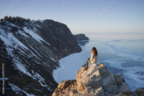 Lake Baikal at winter. Woman sitting on a edge of rock and looking at frozen Baikal lake. Deepest and largest fresh water lake. Olkhon Island, Russia, Siberia.