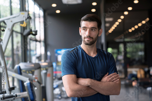 Caucasian man with beard in blue color sportswear standing and putting hands on waist in gym or fitness club.