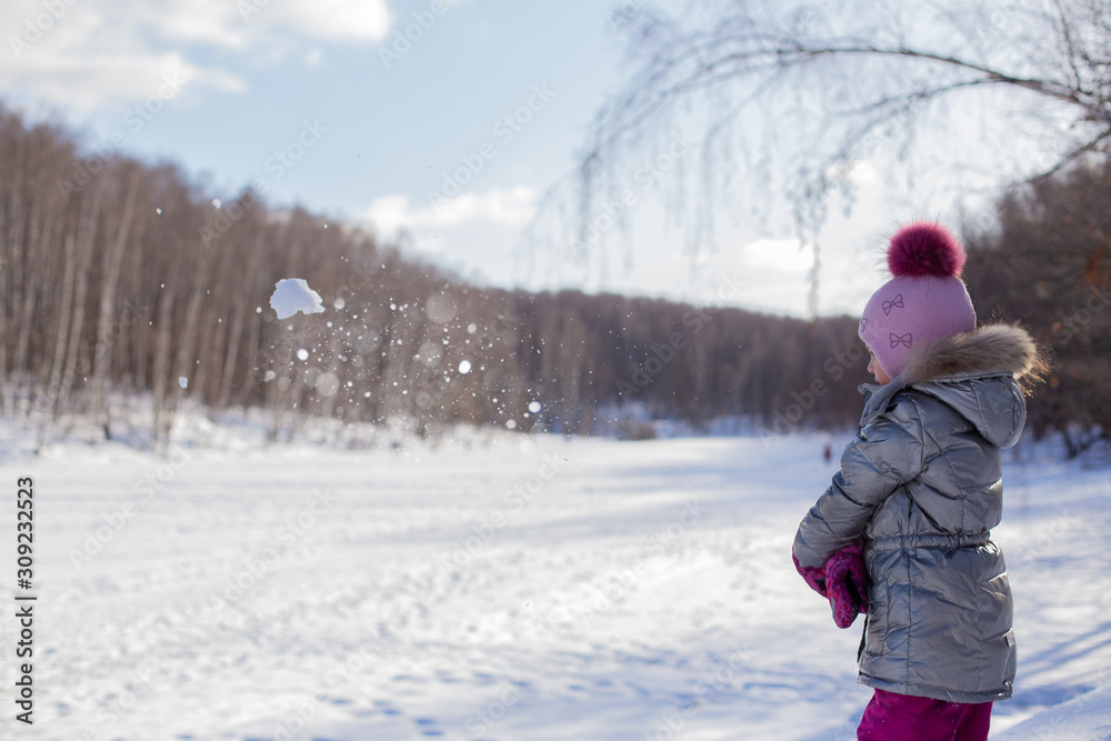 Little girl in a silver jacket and a hat with a pompom walks in a winter park makes and throws large snowballs