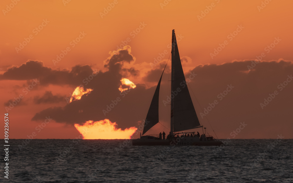 Fototapeta premium a setting sun with a silhouetted sailboat