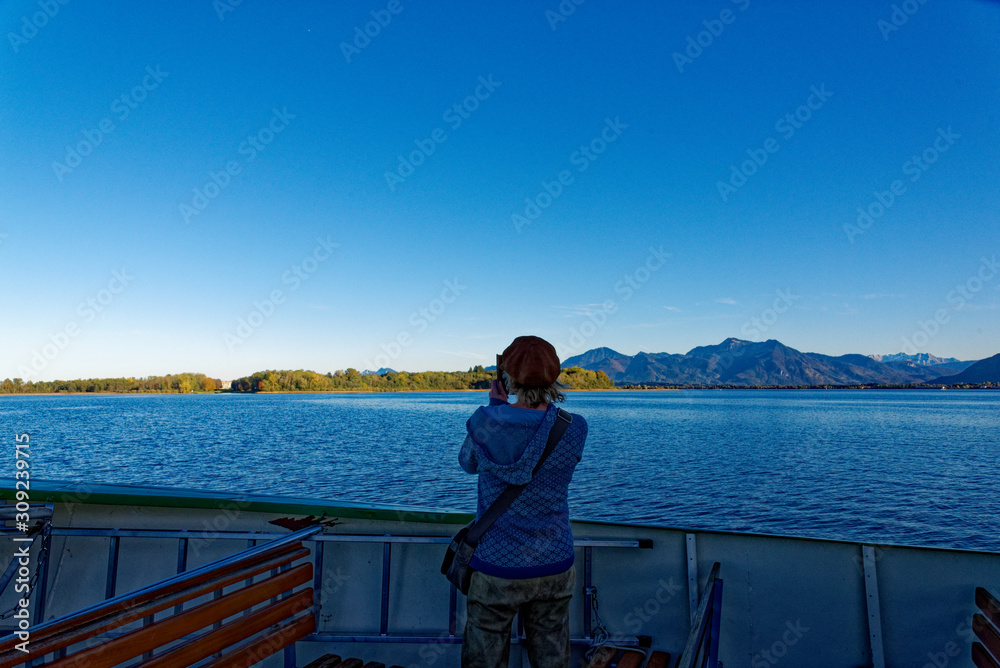 junge Frau steht am Chiemsee und fotografiert die Landschaft im Hintergrund mit Alpen panorama
