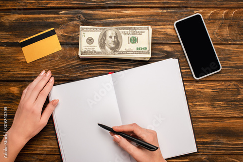 cropped view of businesswoman writing in blank notebook, smartphone, dollar banknotes and credit card on wooden desk
