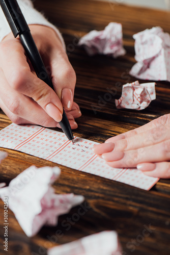 partial view of gambler marking numbers in lottery ticket near crumpled lottery cards on wooden table