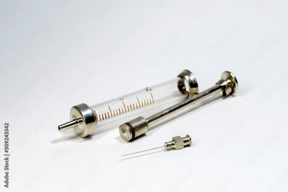 A glass syringe lies on a white tabletop next to a needle. Selective focus. On white background. A glass syringe lies on a white tabletop next to a needle. Selective focus. On white background.