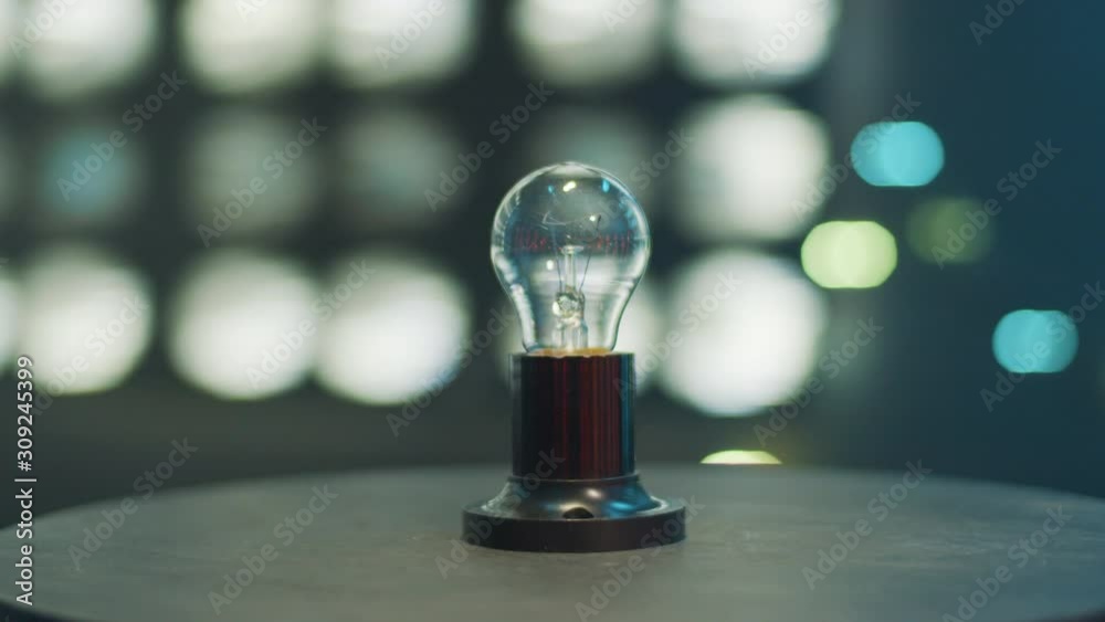 Camera is actively moving, rotating around one switched off bulb lamp placed on dark round-shaped table in room with dark-blue walls and illumination lights.